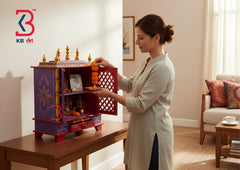 A woman in a light beige kurta stands beside a small decorative altar cabinet on a wooden table in a bright living room. The cabinet is purple with gold and orange accents, open to show framed photos and offerings inside.