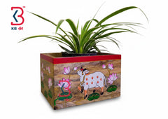 A rectangular hand-painted wooden planter box with a red top rim, featuring a white spotted goat, pink lotus flowers, and green leaves on a brown wood-grain style background. A green spider plant is potted in the wooden planter.