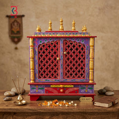 A colorful Indian wooden Shiva mandir altar cabinet with intricate carvings, red lattice doors, and gold finials, displayed on a wooden table with incense, flowers, and a book against a textured beige wall.