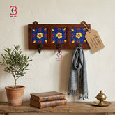 A rustic wooden wall-mounted coat rack with three black hooks and three decorative panels featuring blue, red, and yellow floral patterns. A gray scarf is draped over one hook, and a paper tag hangs from the right hook.