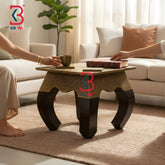 KB ART square antique style brass stool used as a low bedside accent table holding a small decorative white bowl in a sunlit bedroom setting next to stacked magazines.