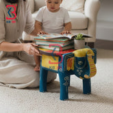 Wooden Elephant Stool KB-5114: A colorful elephant-shaped wooden stool in blue, yellow, and red, styled in a cozy living room setting with a child sitting on it reading