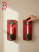 Two red candle holder wall sconces with green candle holders and decorative diamond-shaped tops, each holding a white candle with a flame. A hand is touching the candle on the right.