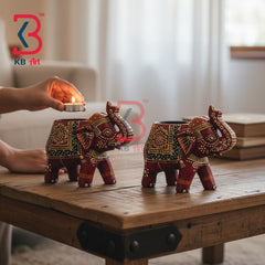 Two decorative elephant-shaped candle holders with intricate red, gold, and green patterns sit on a rustic wooden table in a softly lit room. A hand holds a small burning candle in front of one holder, and a KB Art logo watermark is visible.