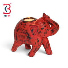 A red elephant-shaped candle holder with a distressed black finish, featuring a built-in tea light cup on its back. The candle is lit, casting a warm glow, and is photographed on a clean white background with a KB Art logo in the top-left corner.