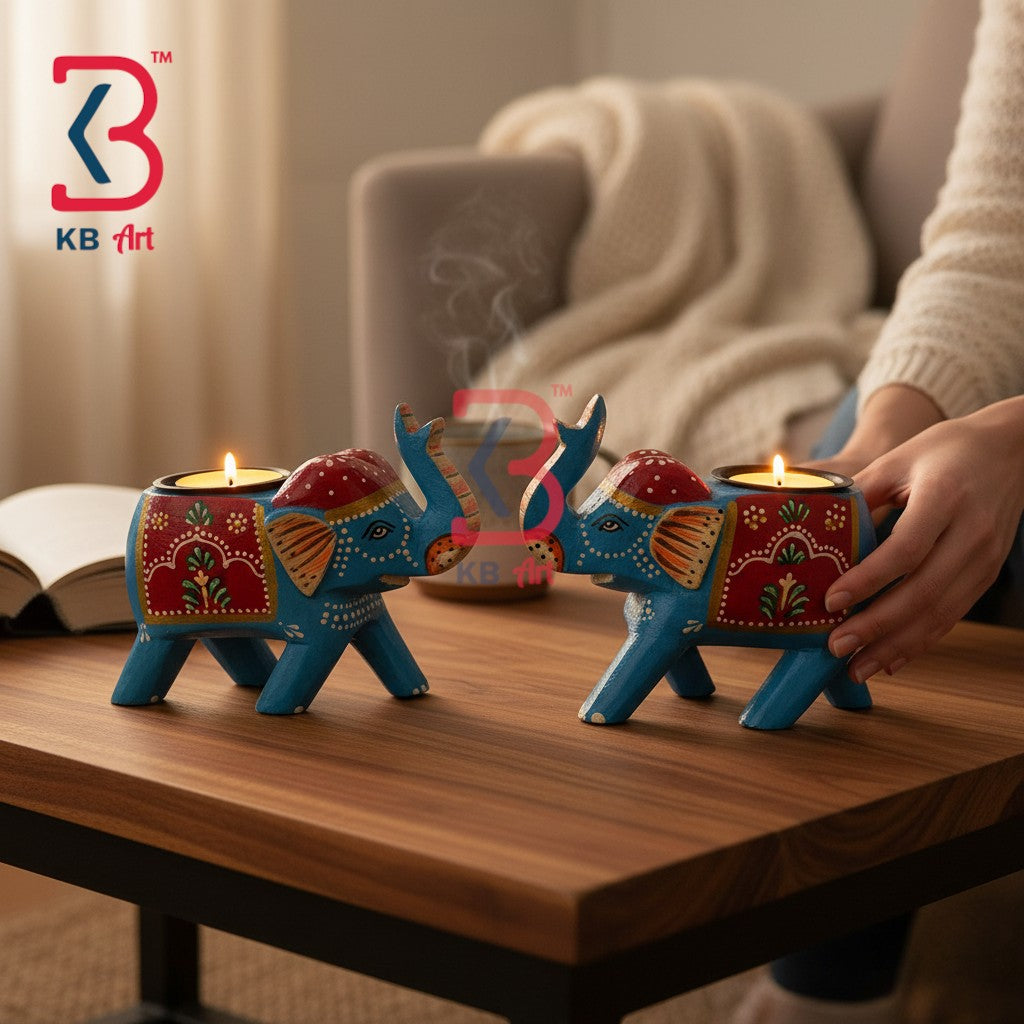 Two decorative elephant-shaped tealight candle holders in blue and red with gold painted details, shown on a wooden table in a cozy indoor setting. A person's hand is adjusting one candle holder, and aKB Art logo is visible in the top left corner.