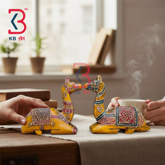 Two small hand-painted camel figurines in yellow with intricate multicolor patterns, shown on a table with hands holding them near a steaming cup in a softly lit indoor setting. KB Art logo is visible in the top left.