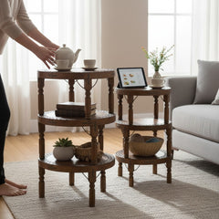 Two round wooden three-tier side tables in a bright living room. One table is styled with a teapot, cups, books, and a tablet, while the other is styled with a small plant, vase, and basket.