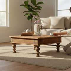 A rustic wooden coffee table with turned legs sits on a light beige rug in a bright living room. A person is partially visible on the right, holding a cup near the table, with a sofa, plant, and windows in the background.