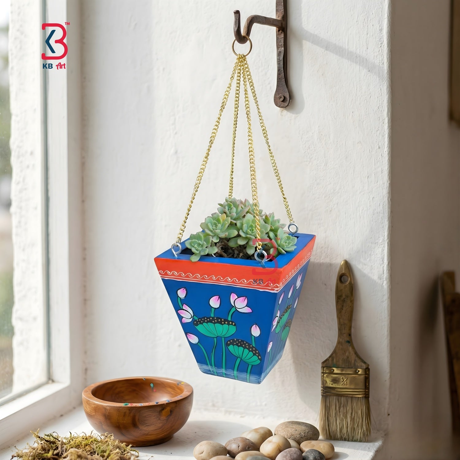 Hanging planter with floral design on a windowsill with a wooden bowl and brush.