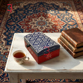 A decorative rectangular tea tin with a blue-and-white floral lid and red base sits on a white table beside a cup of tea and a stack of old books, set on a patterned rug.