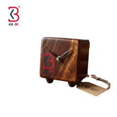 A small wooden clock with a square shape, featuring a dark brown wood grain finish, bronze-toned hands, and a red and blue '3' logo on the front. It has small feet and a hanging tag attached to a cord, set against a plain white background with a 'KB Art' logo in the top-left corner.