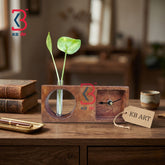 A rustic wooden desk clock and vase set on a wooden table, featuring a rectangular wood block with a circular cutout holding a green plant stem and a small square compartment with a clock face. A KB Art branded tag hangs from the front, with books and a cup in a softly blurred background.