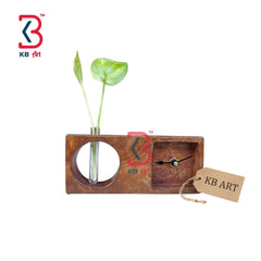 A rectangular wooden wall clock with a circular cutout holding a green plant stem and a small inset clock face. The wood has a warm brown finish, and a small paper tag reading KB ART hangs from the clock.