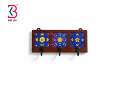 A rectangular wooden wall rack with three hanging hooks and three blue decorative tile panels featuring yellow and red floral patterns, shown against a plain white background.