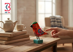 A colorful hand-painted wooden bird figurine on a small teal base, displayed on a wooden table next to a teapot and books in a cozy living room setting, with a human hand pointing toward it.