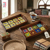 Two decorative wooden serving trays with colorful mosaic tile patterns, displayed on a rustic wooden table alongside a teacup, books, a woven plate, and a small wooden box. The KB Art logo is visible in the top-left corner.