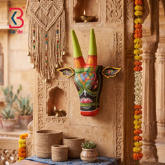 A hand-painted traditional Indian wooden wall hanging depicting a decorated bull head with green horns, displayed on a carved stone pillar in a warm, rustic indoor setting with decorative garlands and small plants.