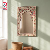 An ornate rectangular wall mirror with an arched center and a hand-painted floral border, mounted on a textured sage green wall. The mirror is styled in a room setting above a wooden table with a woven basket and books, with soft sunlight casting shadows on the wall.