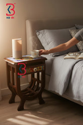 A dark brown wooden nightstand beside a bed, styled with a white cylindrical lamp and a steaming cup on a colorful mat. A person's arm reaches toward the cup.