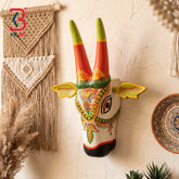 Multicolor hand-painted wooden cow head wall hanging with orange horns, yellow sunburst and floral pendant design on white background