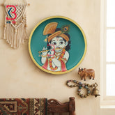 A round decorative wall plate featuring an illustration of Lord Krishna, displayed on a beige wall alongside a macrame wall hanging, a small cow figurine, and a beaded bell.