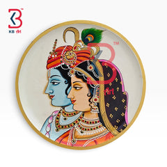 A circular decorative wall art print featuring a traditional Indian illustration of Lord Krishna and Radha in profile, with a gold-toned border and a white background. A red and blue KB Art logo watermark is visible in the top-left corner.