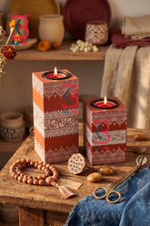 Two decorative rectangular candle holders with red and orange geometric patterns, holding lit tealight candles, displayed on a rustic wooden table with a beaded necklace, scissors, and dried flowers in a warm, bohemian setting.