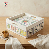 A decorative rectangular serving tray with cutout handles and small front drawers, decorated with colorful geometric and floral tile-style patterns on a light wood table. A brass cup, folded cream cloth, and a KB Art tag are also visible in the styled setting.