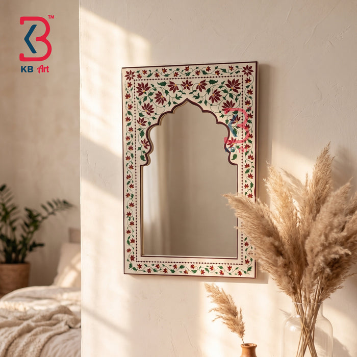 An arched rectangular wall mirror with an ornate floral border in red, green, and cream, hanging on a textured beige wall in a softly lit bedroom. Decorative dried pampas grass and a bed are visible in the background.