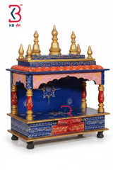 A hand-painted Indian miniature Hare Krishna gopuram (temple tower) with a blue and gold painted structure, featuring ornate gold finials, red and gold decorative panels, and a blue arched niche. The KB Art logo is visible in the top-left corner.