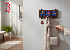 A wall-mounted wooden coat rack with three blue floral tile panels and hanging keys, scarf, and a beige handbag, shown in a bright interior setting. A person's hand is visible holding the bag.