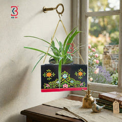 Decorative hanging planter with floral patterns on a wooden surface near a window with garden view.