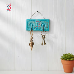A turquoise blue rectangular wall-mounted key holder with carved floral details and two hooks, shown hanging on a white paneled wall. Two vintage-style keys are attached, along with a rope key bag.
