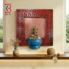 A decorative red wooden wall plaque with carved circular patterns, displayed on a shelf in a home setting next to a small vase and a spool of thread, featuring a brass statue of Lord Ganesha on a blue pedestal.