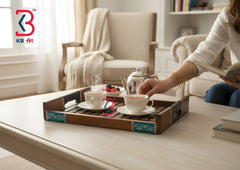 A decorative wooden serving tray with teal accents holds two white teacups, a small silver teapot, and berries on a light-colored table in a cozy living room setting. A person's arm is visible reaching toward the tray.