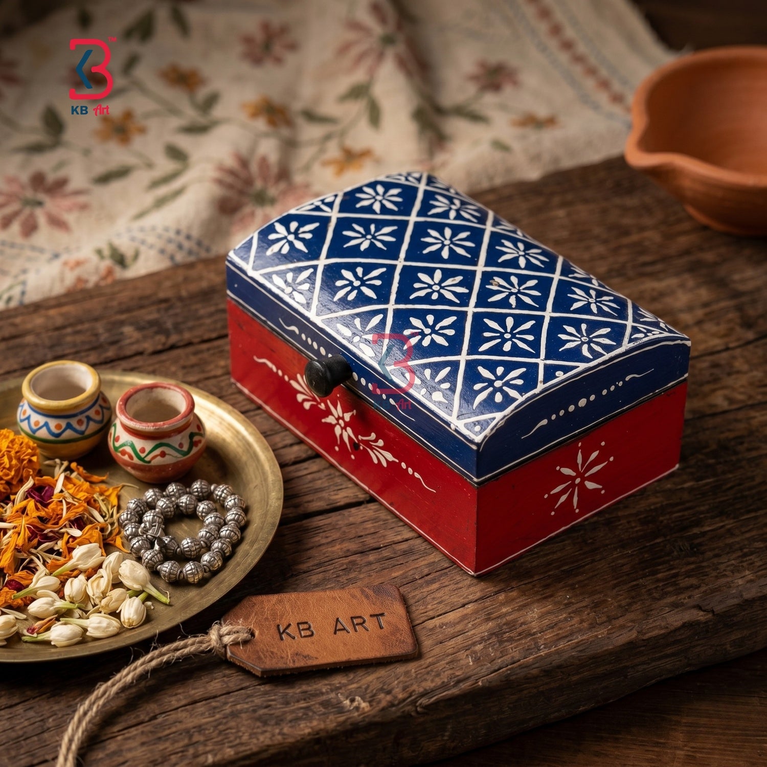 A decorative rectangular metal box with a blue and white floral pattern on top and a red base with white floral motifs, placed on a rustic wooden surface with small decorative bowls, dried flowers, and a hanging tag reading KB ART.
