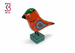 A hand-painted wooden bird figurine in vibrant red-orange with green, black, and blue decorative details, mounted on a teal square base against a white background with a KB Art logo in the top-left corner.