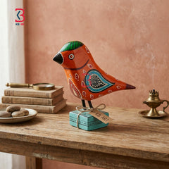 A decorative hand-painted bird figurine in orange, green, black, and turquoise stands on a small turquoise gift box tied with a ribbon. The scene is styled on a rustic wooden table with books, a brass magnifying glass, a small dish with stones, and a brass lamp against a warm terracotta wall.