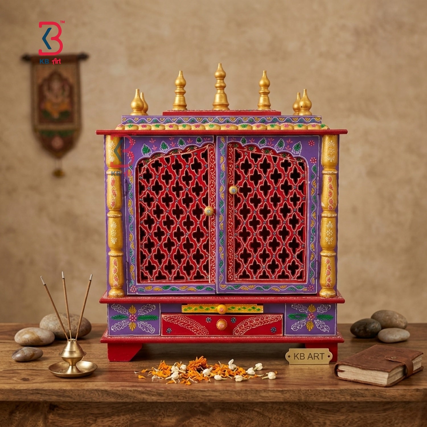 A colorful Indian wooden Shiva mandir altar cabinet with intricate carvings, red lattice doors, and gold finials, displayed on a wooden table with incense, flowers, and a book against a textured beige wall.