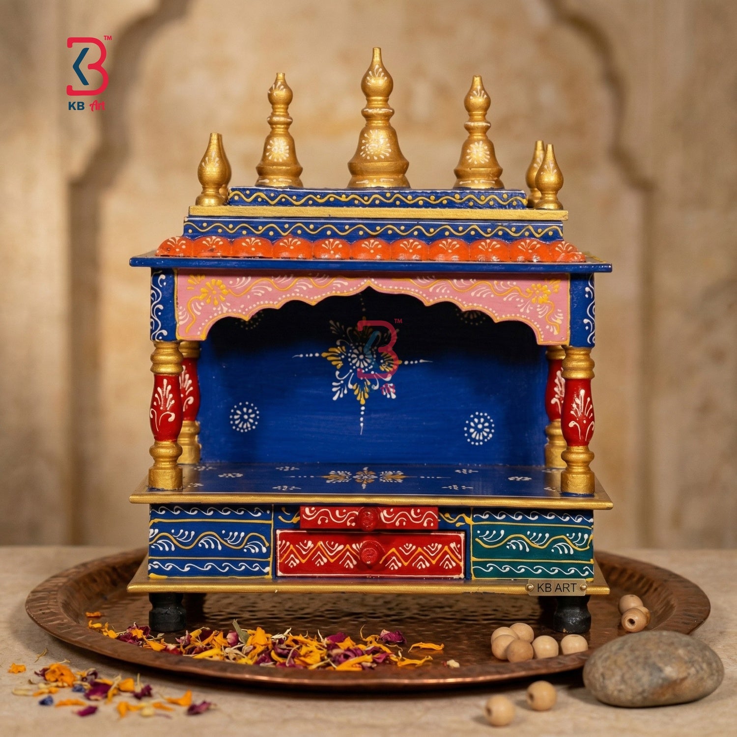 A small decorative wooden altar or holi temple with a blue body, gold finials, red drawer panels, and pink upper trim, displayed on a tray with dried flowers and stones against a warm stone background.