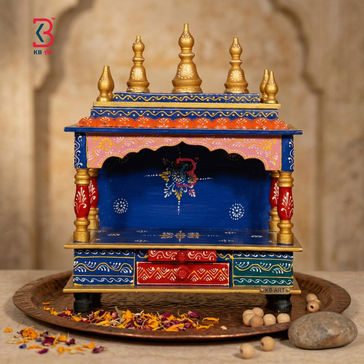 A small decorative wooden altar or holi temple with a blue body, gold finials, red drawer panels, and pink upper trim, displayed on a tray with dried flowers and stones against a warm stone background.