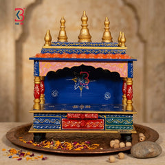 A small decorative wooden altar or holi temple with a blue body, gold finials, red drawer panels, and pink upper trim, displayed on a tray with dried flowers and stones against a warm stone background.