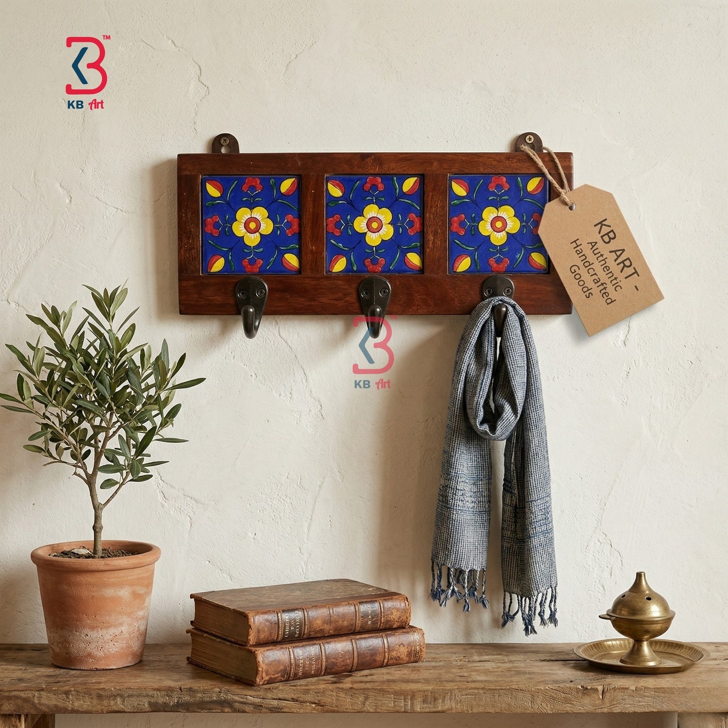 A rustic wooden wall-mounted coat rack with three black hooks and three decorative panels featuring blue, red, and yellow floral patterns. A gray scarf is draped over one hook, and a paper tag hangs from the right hook.