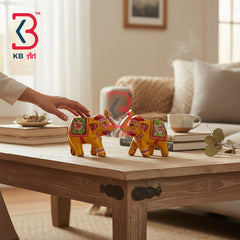 Two small decorative elephant figurines with yellow bodies and colorful floral saddles are placed on a wooden coffee table in a living room. A person's hand reaches toward one elephant, and books, a cup, and a sofa are visible in the background.