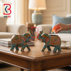 Two decorative elephant-shaped tealight candle holders with intricate multicolor patterns in turquoise, gold, pink, and green, shown on a wooden coffee table in a living room setting. A hand is lighting the candle on the right, with a KB Art logo watermark in the top left corner.