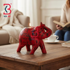 A red elephant-shaped tea light candle holder with a distressed finish sits on a wooden coffee table in a cozy living room setting. A person is visible in the background reading a book, and a KB Art logo watermark is present in the upper left corner.