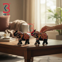 Two decorative elephant figurines with colorful traditional patterns on black bodies, displayed on a wooden table in a softly lit living room. A hand is adjusting one figurine, and a KB art watermark is visible.