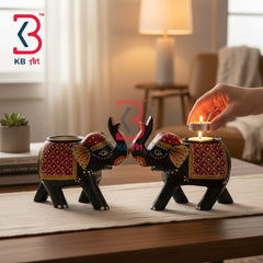 Two decorative elephant-shaped candle holders on a wooden table in a warmly lit living room. A human hand is lighting a small tea light candle placed in one holder.