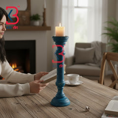 A blue wooden candlestick holder with a lit candle sits on a rustic wooden table in a cozy living room setting. A woman is seated at the table reading a book, with a fireplace, sofa, and cup in the background.